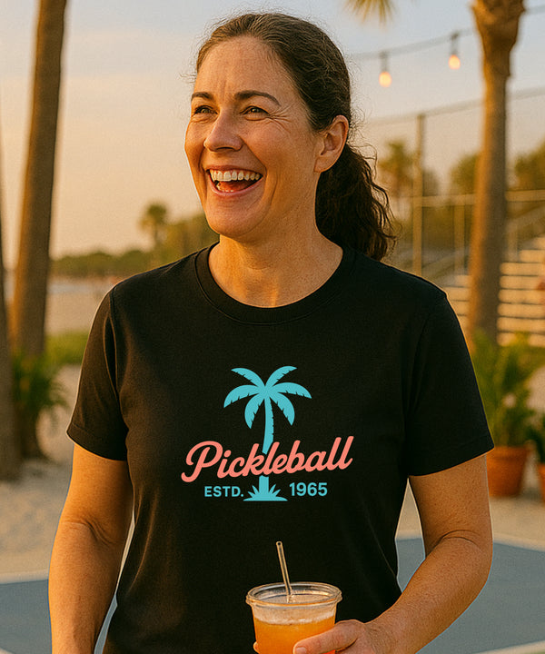 Woman wearing a black t-shirt with a palm tree and 'Pickleball' design, holding a drink outdoors.