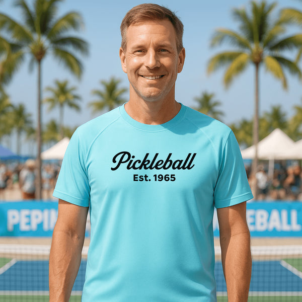 Man wearing a blue 'Pickleball Est. 1965' t-shirt on a pickleball court with palm trees in the background.