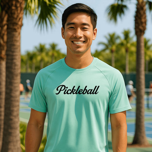Man wearing a teal 'Pickleball' shirt standing on a pickleball court with palm trees in the background.